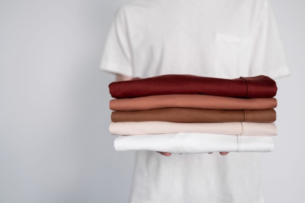 A person holding a stack of neatly folded shirts in earthy tones against a soft white background.